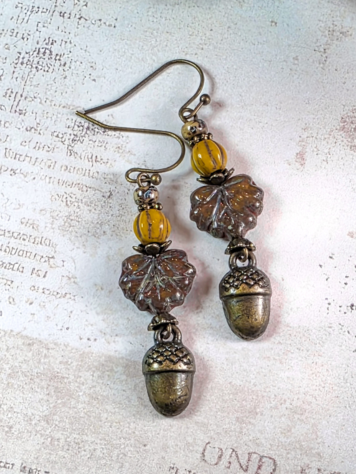 Acorn and leaf dangle earrings with antique bronze metal, amber-yellow beads, and brown translucent glass elements, displayed on a textured off-white background with faint script.