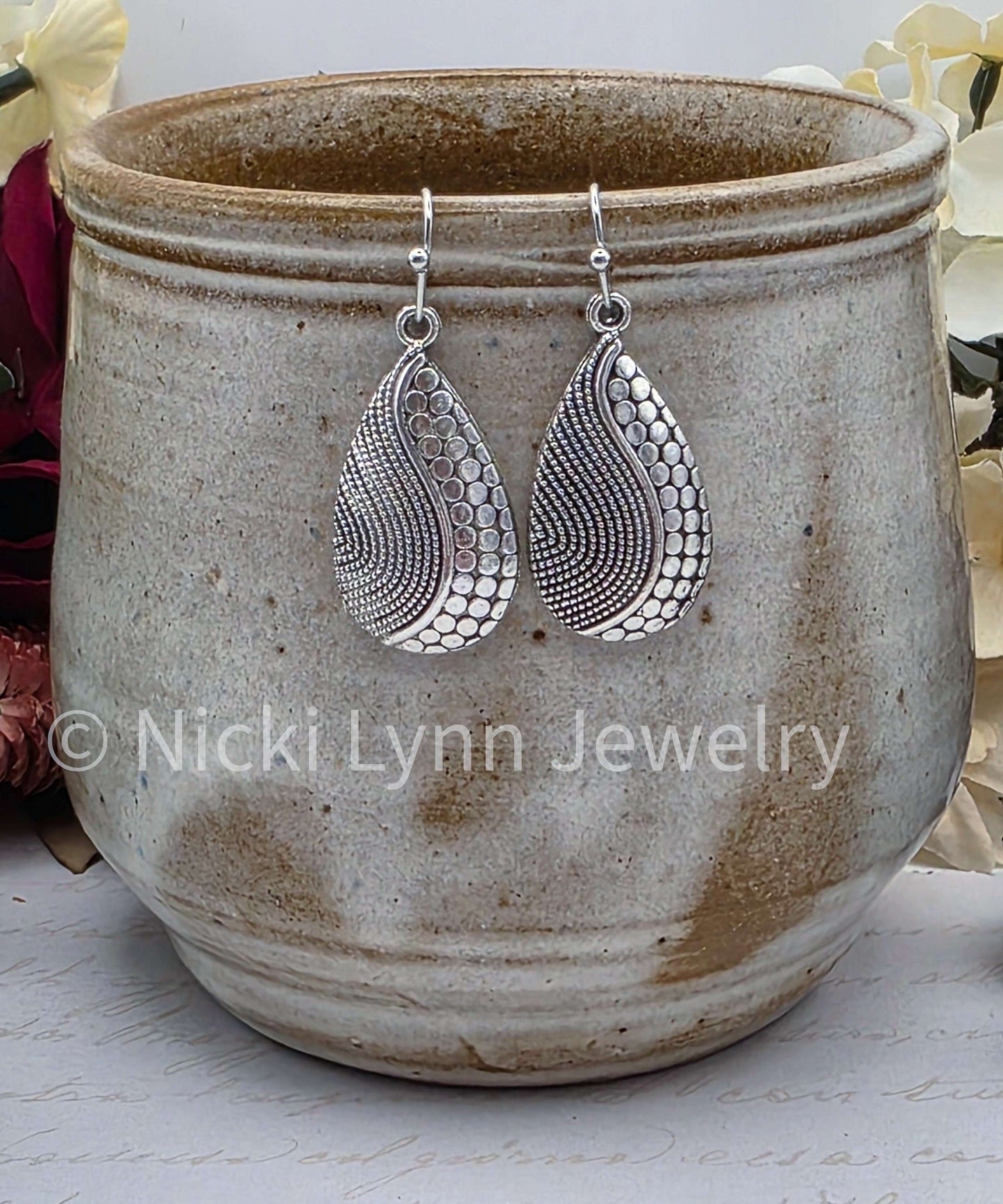 May include: A pair of silver teardrop earrings. Each earring features a detailed pattern of small circles and lines. The earrings are displayed against a neutral background, showcasing their intricate design and craftsmanship. The earrings are a jewelry item. Nicki Lynn Jewelry