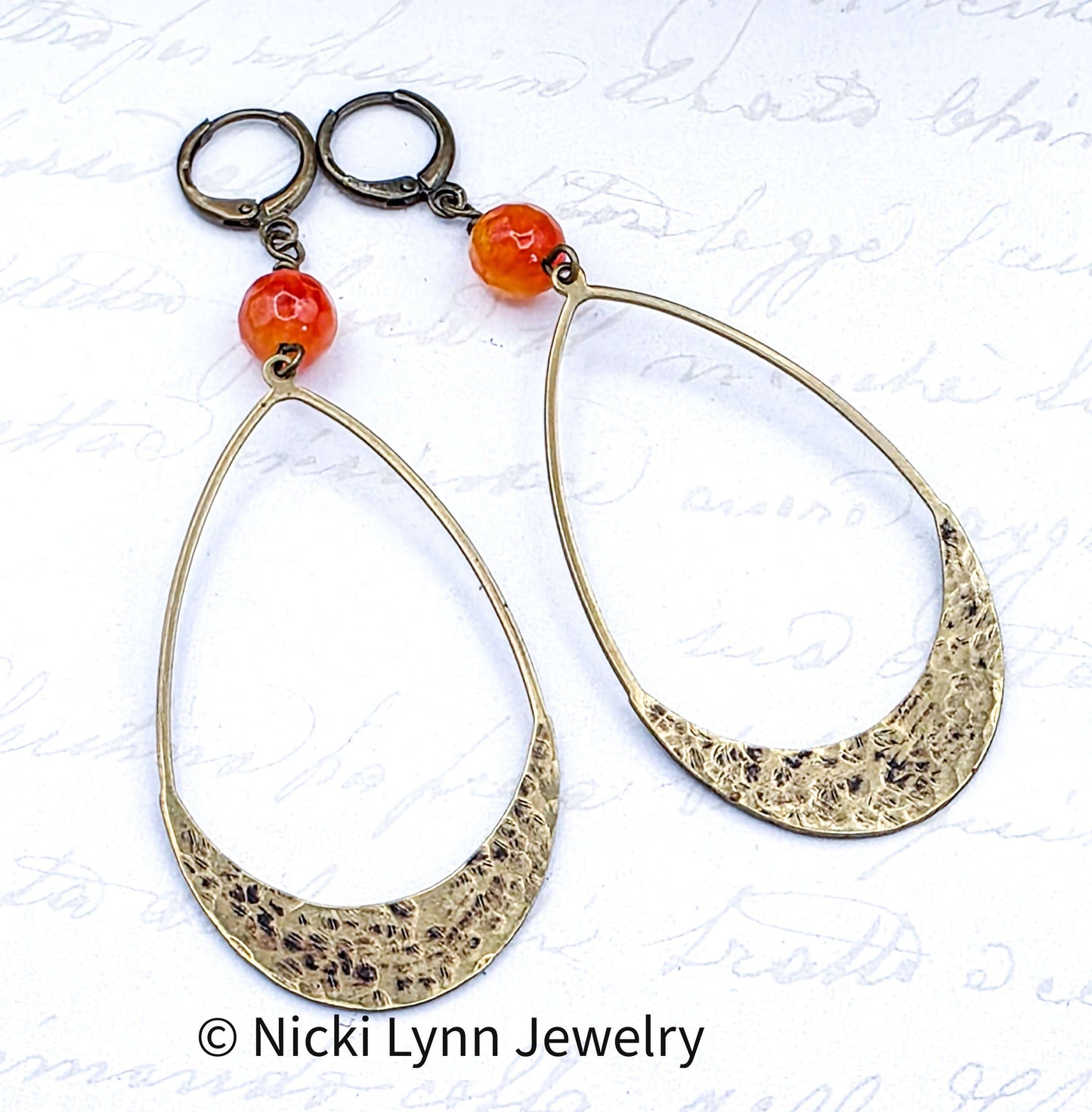 Gold hoop earrings with orange beads on a textured white background, featuring Nicki Lynn Jewelry branding.
