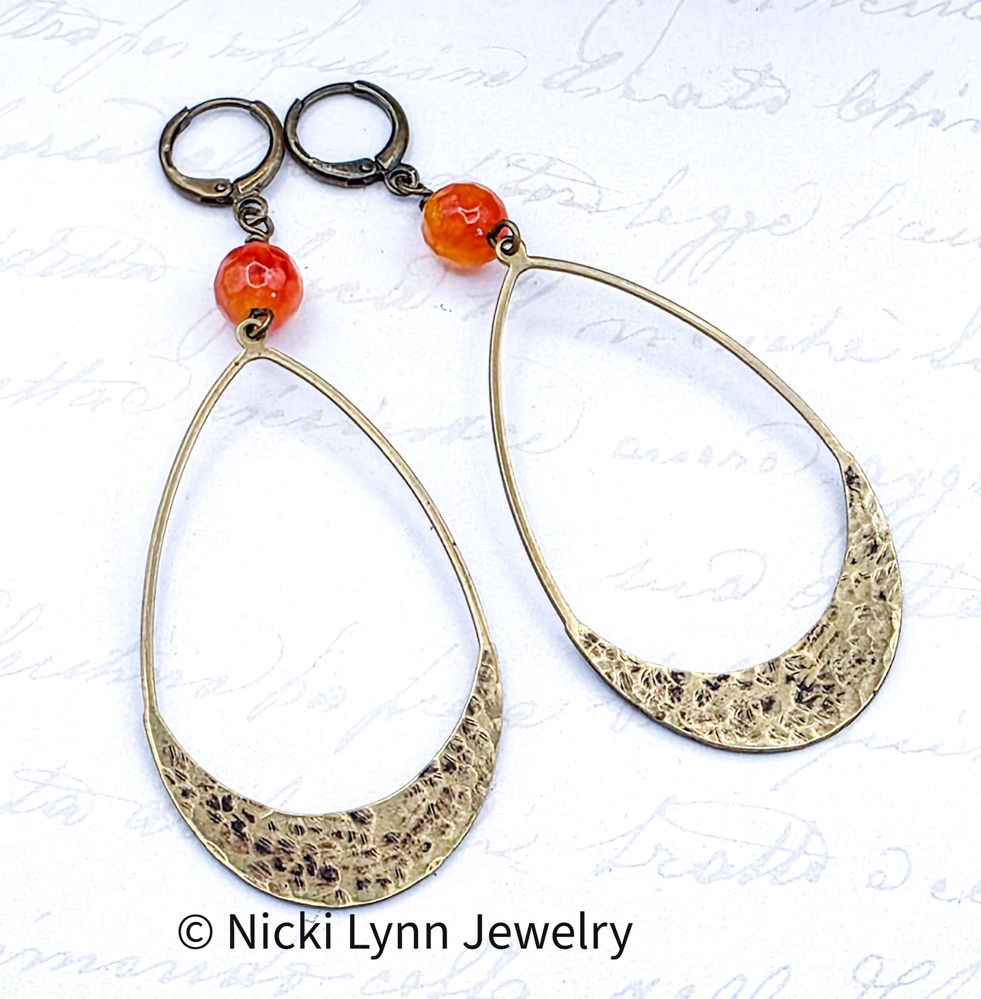 Gold hoop earrings with orange beads on a textured white background, featuring Nicki Lynn Jewelry branding.
