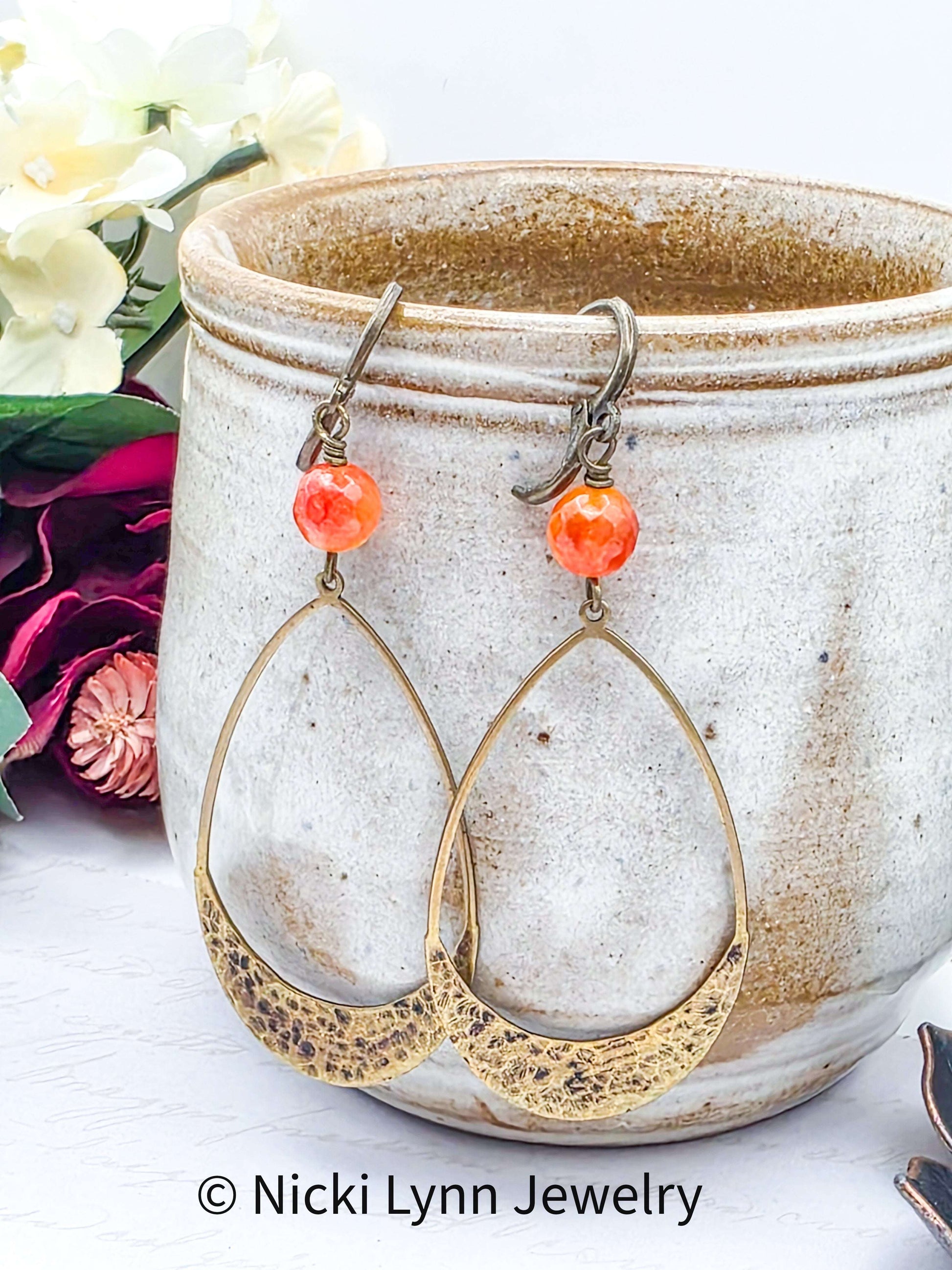 Gold hoop earrings with orange beads on a ceramic container with flowers in the background, branded 'Nicki Lynn Jewelry'.
