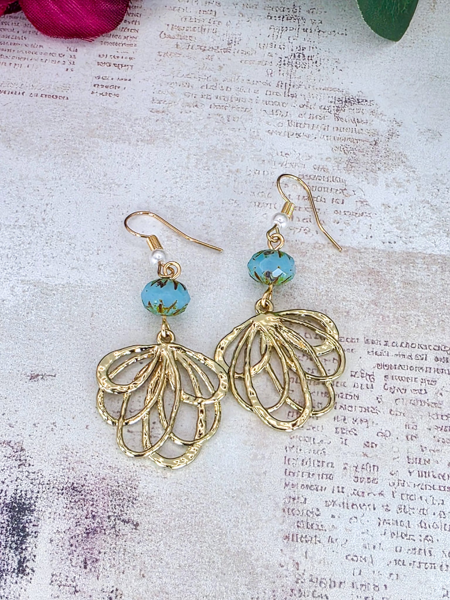 Gold-toned dangle earrings with oval cutout charms and light blue faceted beads, displayed on a textured vintage-style paper background.