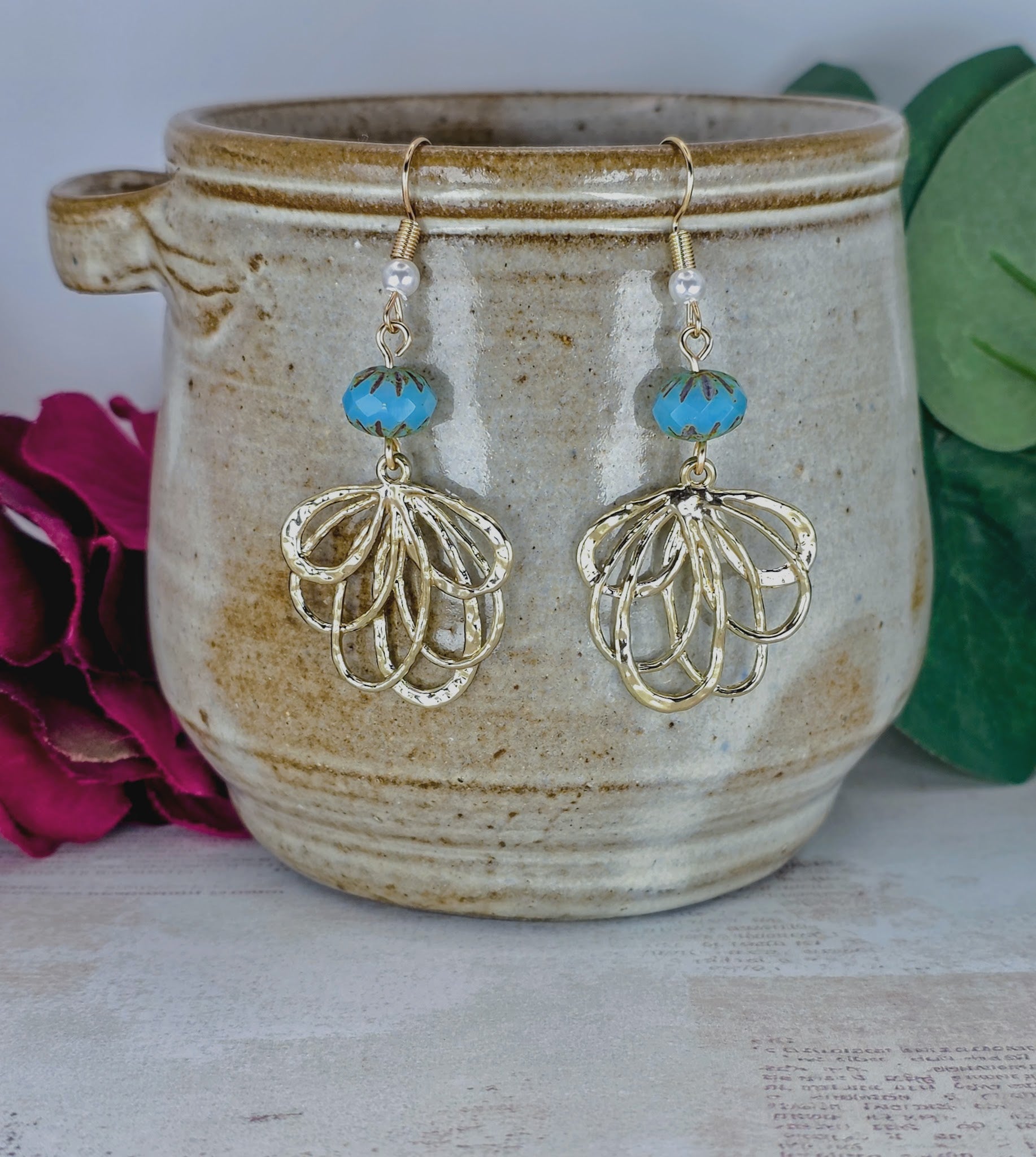 A pair of bohemian-style dangle earrings with gold filigree leaf charms, white beads, and turquoise blue beads, hanging in front of a rustic beige ceramic pot with pink and green decorative props.