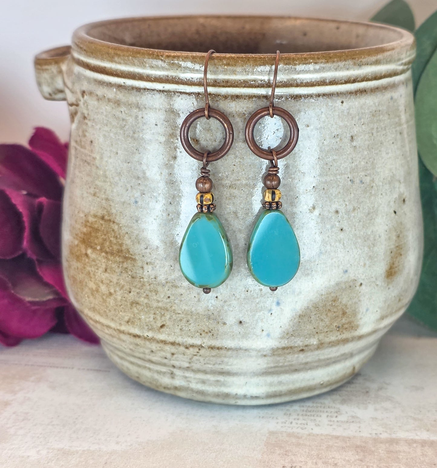 A pair of handmade drop earrings with turquoise teardrop beads and copper-toned metal rings, hanging from a rustic beige ceramic pot with decorative flowers in the background.