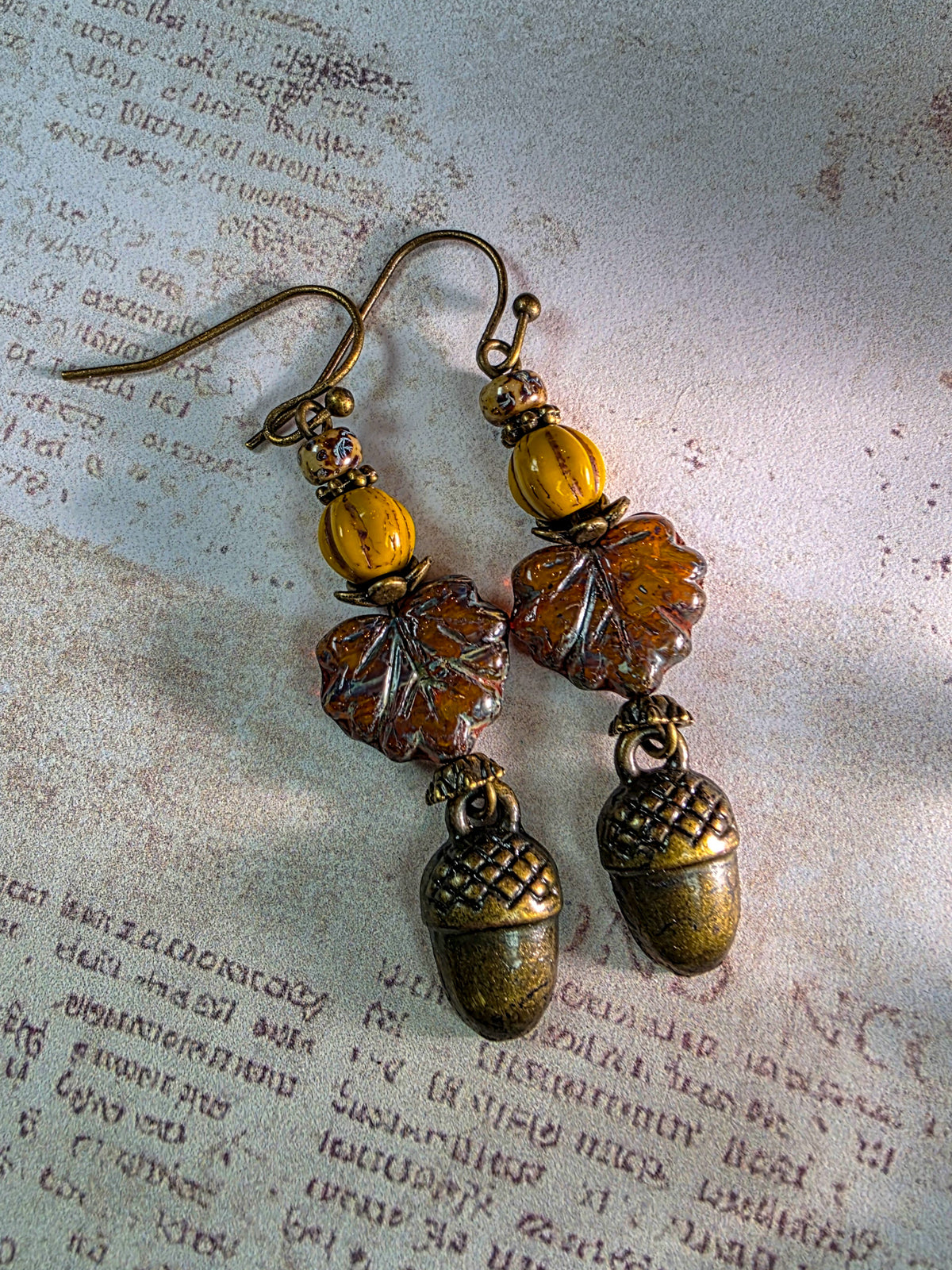 A pair of dangling earrings with antique bronze-toned metal hooks and acorn charms, featuring amber and translucent brown leaf-shaped beads, displayed on a textured vintage book page.