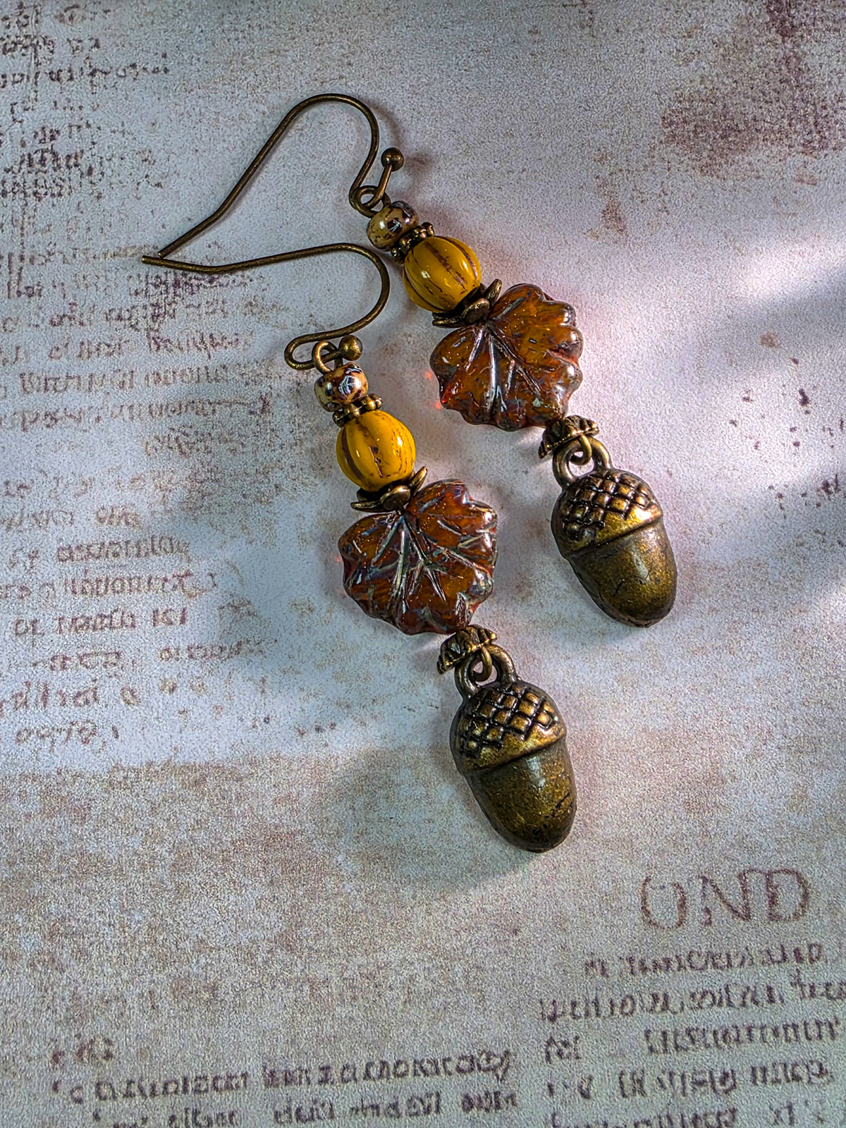 A pair of dangling earrings with antique bronze acorns, amber-brown carved leaf charms, and round amber beads, displayed on a textured, vintage-style beige paper background.