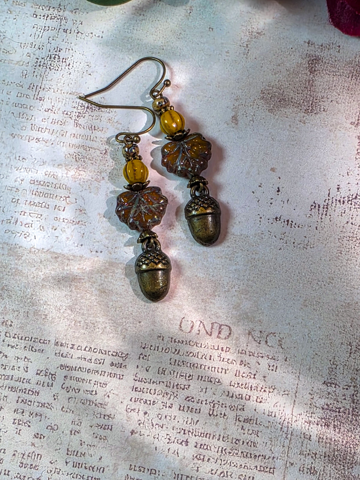 A pair of handmade dangle earrings featuring antique bronze acorns, bronze leaves, amber-yellow beads, and silver-toned hooks, displayed on a textured, vintage-style paper background.
