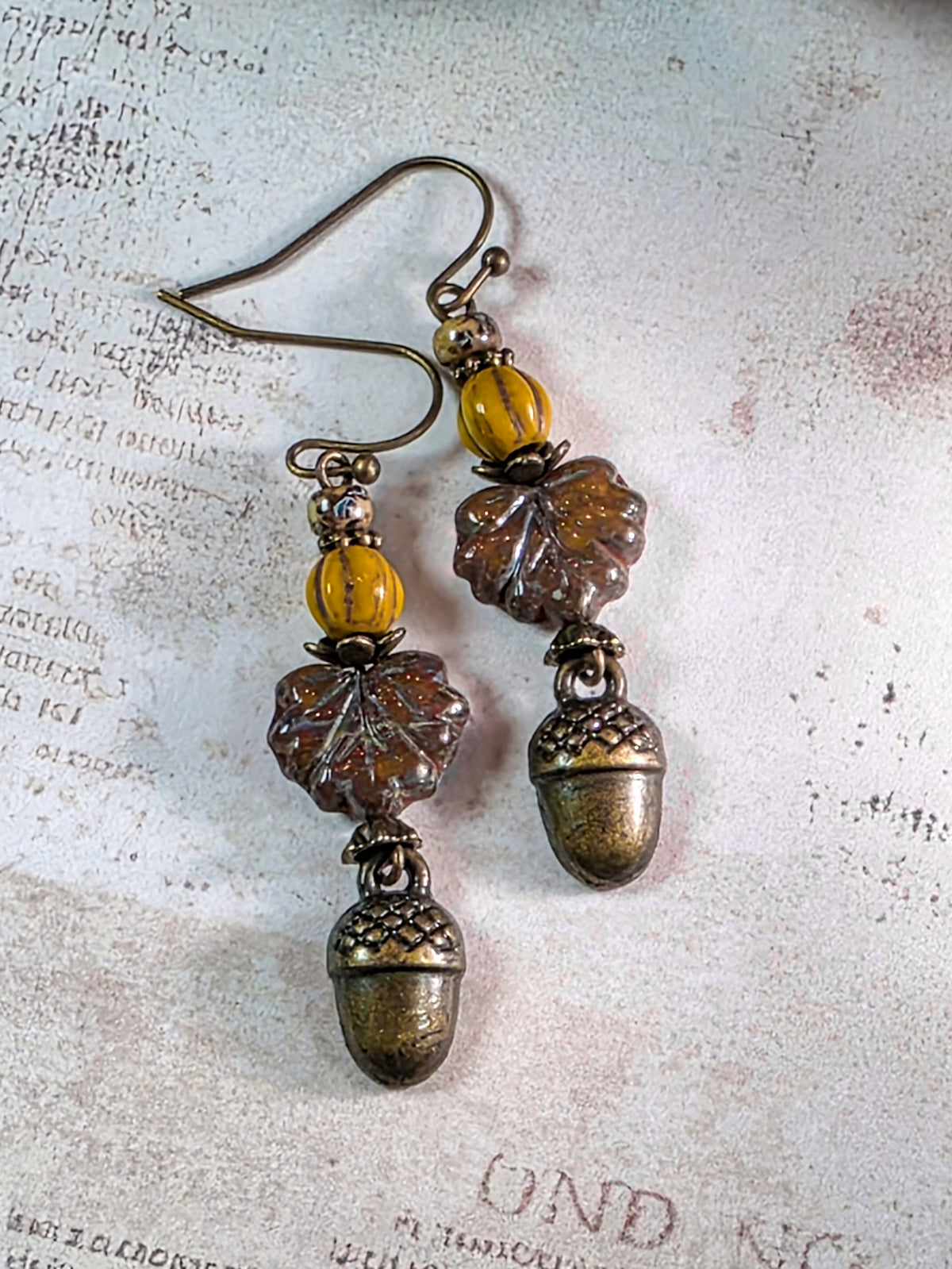 A pair of dangling acorn earrings with textured brown leaf charms, amber-yellow beads, and antiqued silver-tone metal hooks, displayed on a vintage-style paper background.
