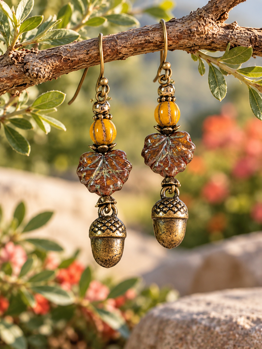 A pair of dangling earrings hanging from a tree branch outdoors. Each earring has an antiqued gold-tone fishhook, a round amber-yellow bead, a textured leaf-shaped charm in brown and amber tones, and a matching acorn pendant at the bottom.